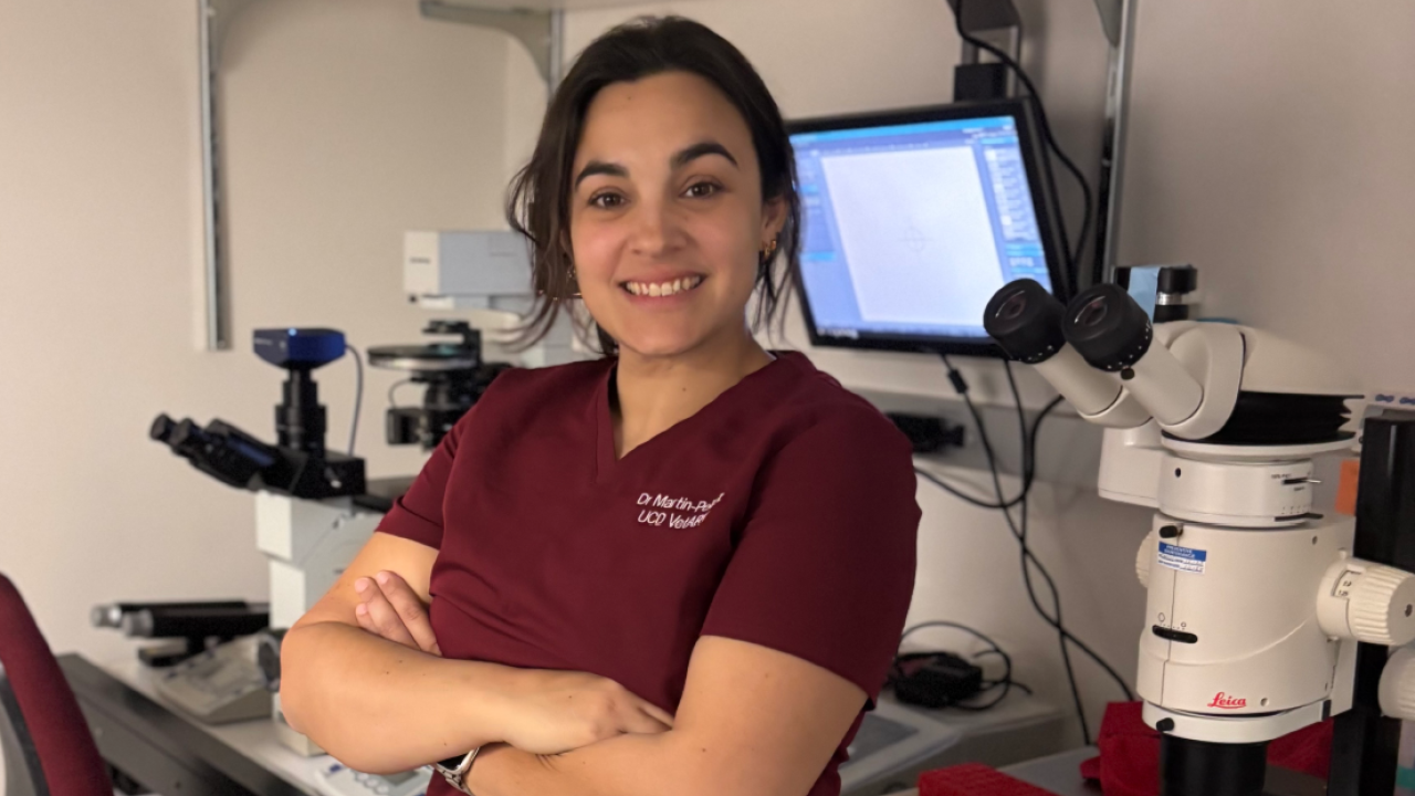 Picture of Dr. Martin-Pelaez in maroon scrubs standing in front of a microscope in the laboratory.